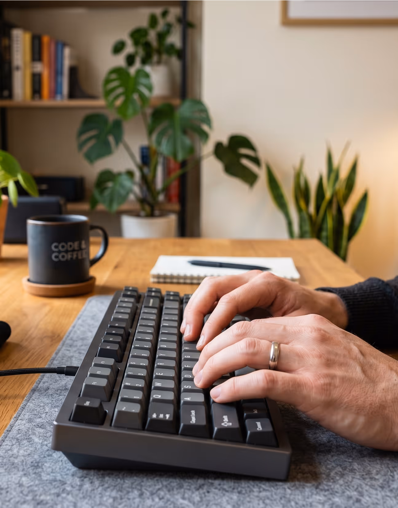 Hands typing on a mechanical keyboard on a desk with plants, a notebook, and a coffee mug nearby.