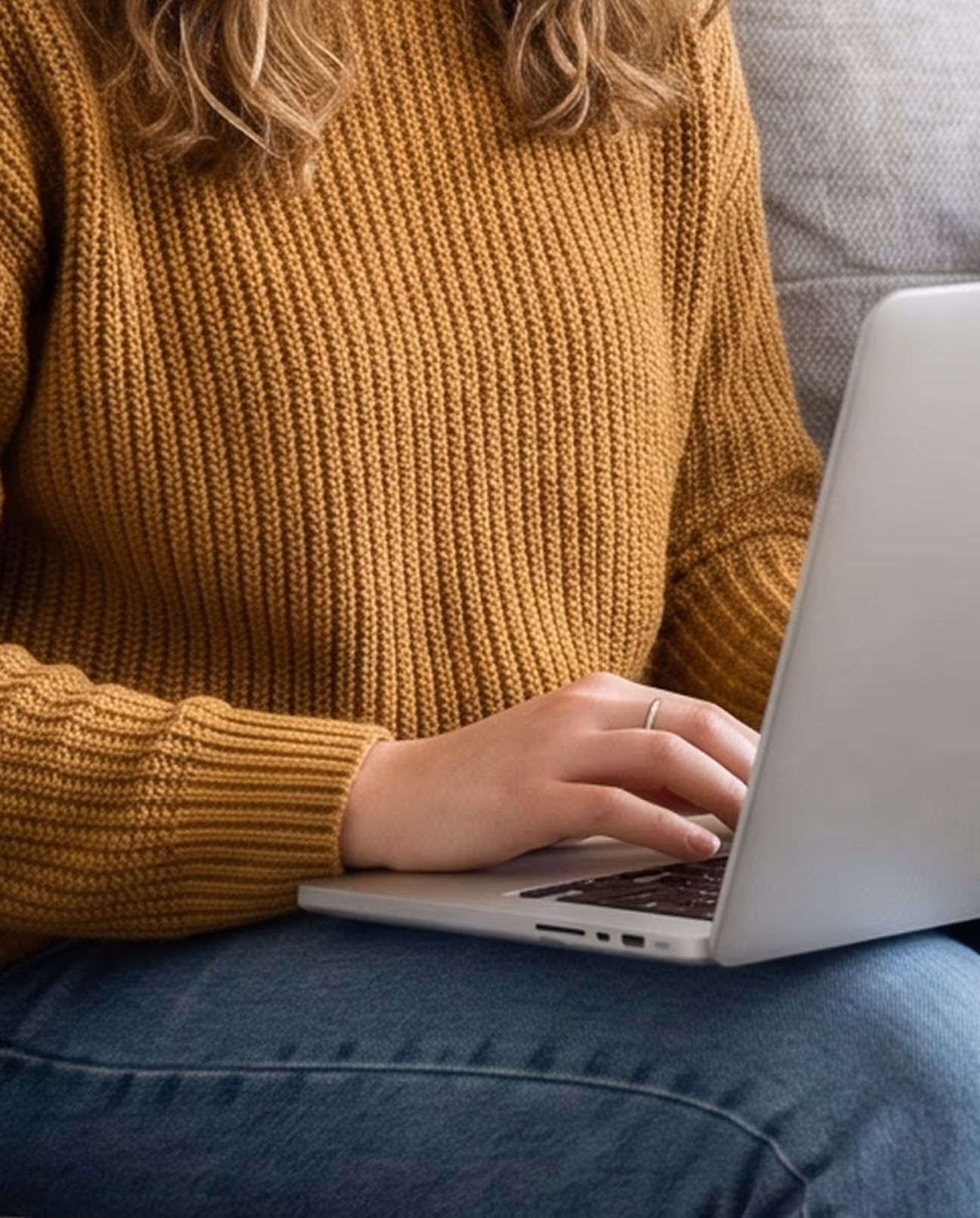 Person wearing a brown sweater and blue jeans typing on a silver laptop on their lap.