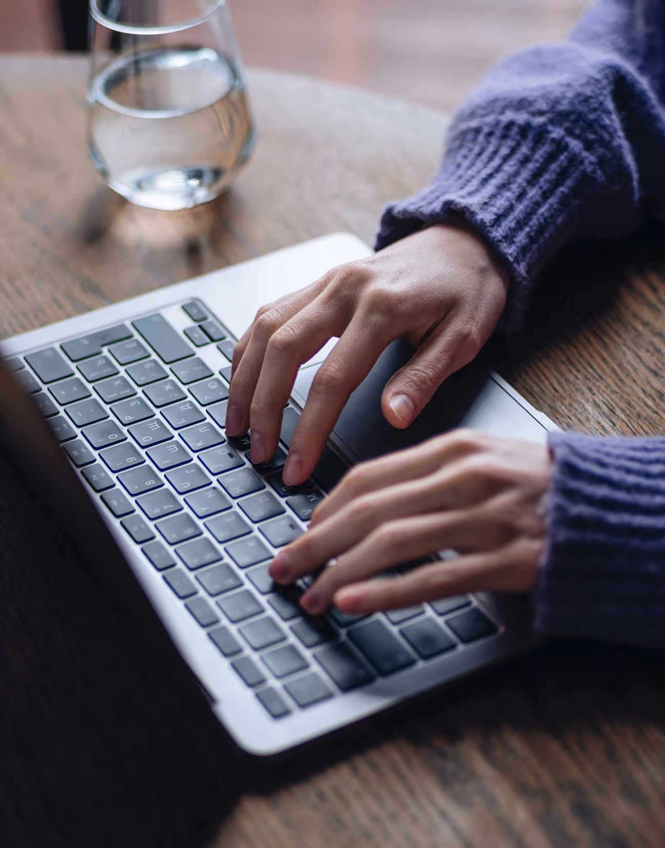 Mains tapant sur un clavier d’ordinateur portable posé sur une table en bois, avec un verre d’eau à côté.