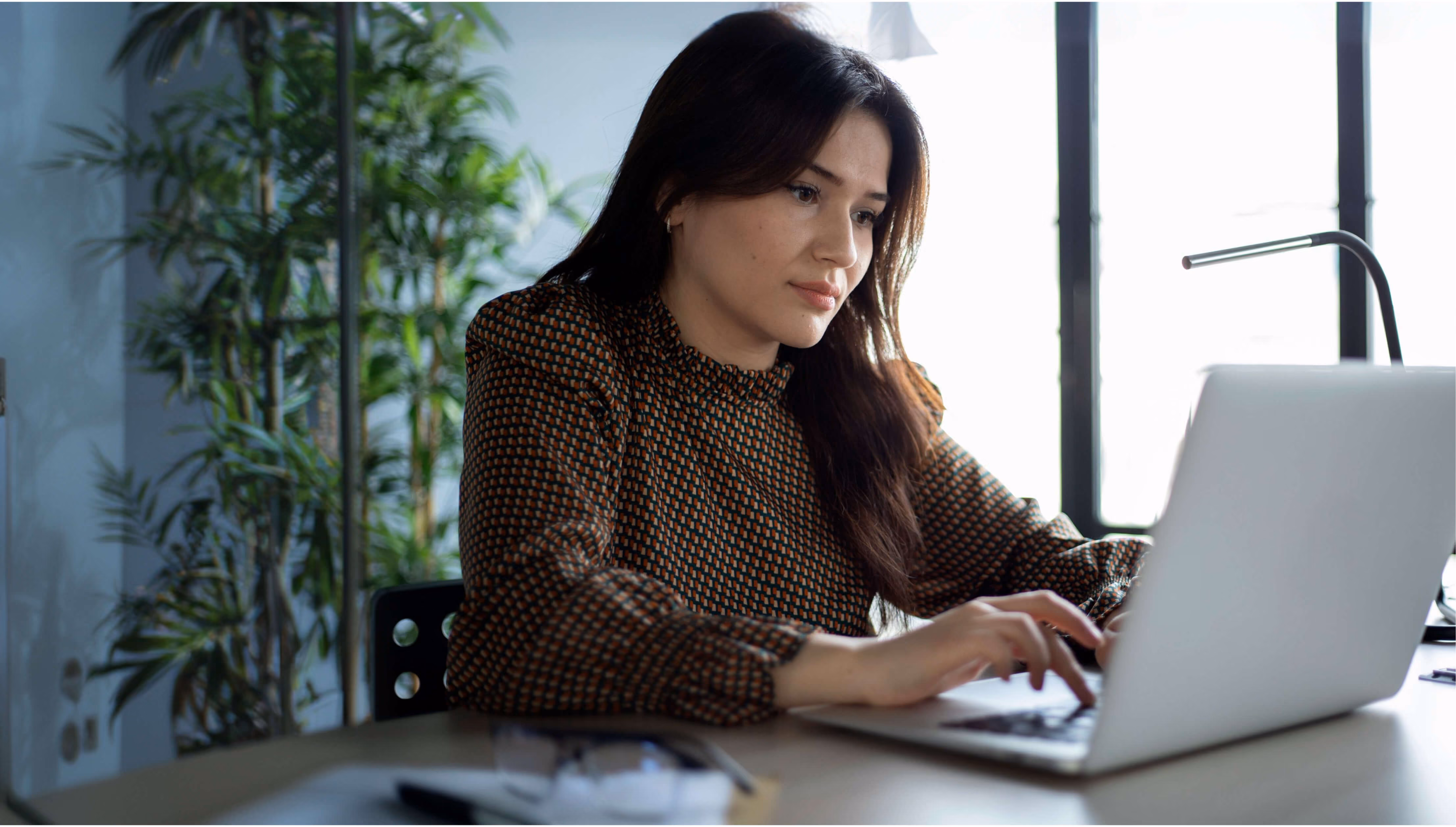 Femme concentrée utilisant un ordinateur portable à une table près d'une grande fenêtre avec une plante verte.