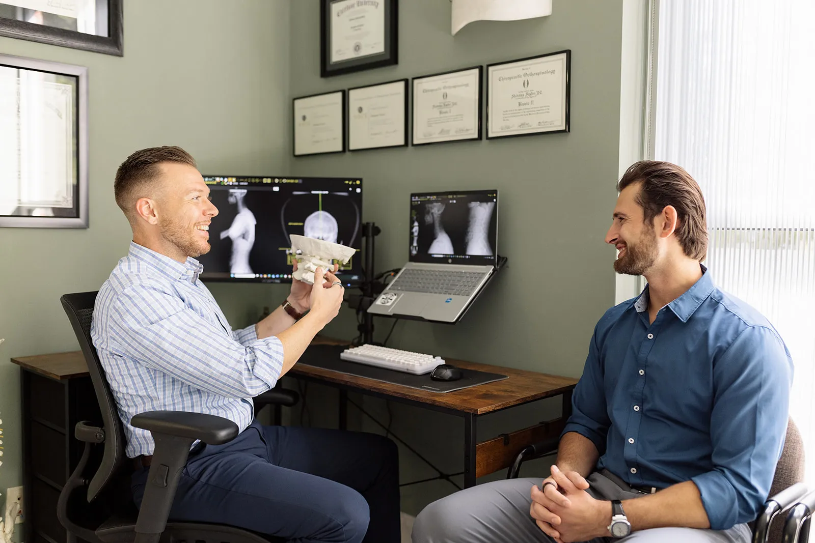 Doctor showing a spine model to a smiling patient in an office with spine X-rays on screens.