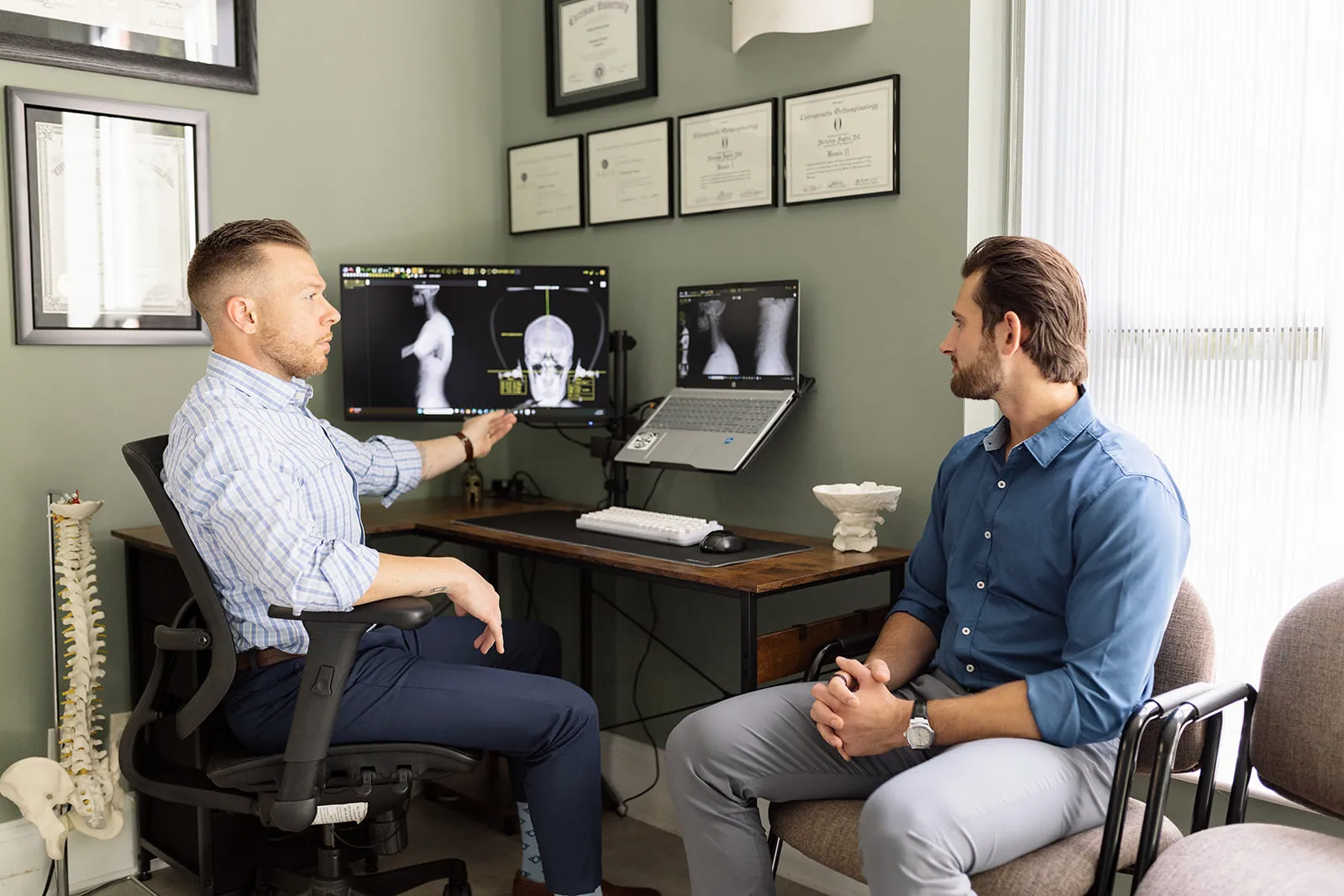 Doctor explaining spine X-rays to a patient in a medical office with diplomas on the wall.