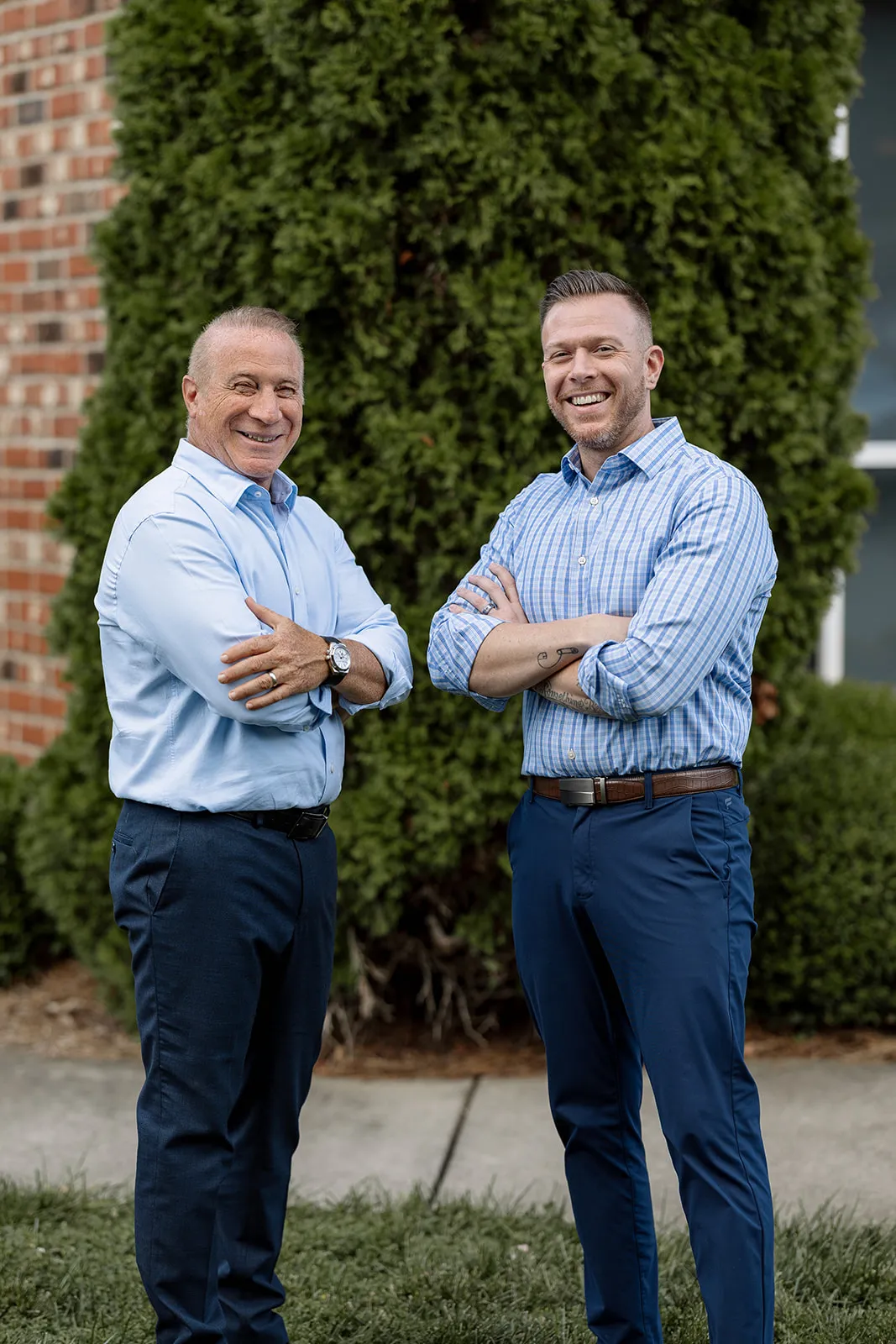 Two men standing outdoors with arms crossed, smiling, wearing blue shirts and navy pants.