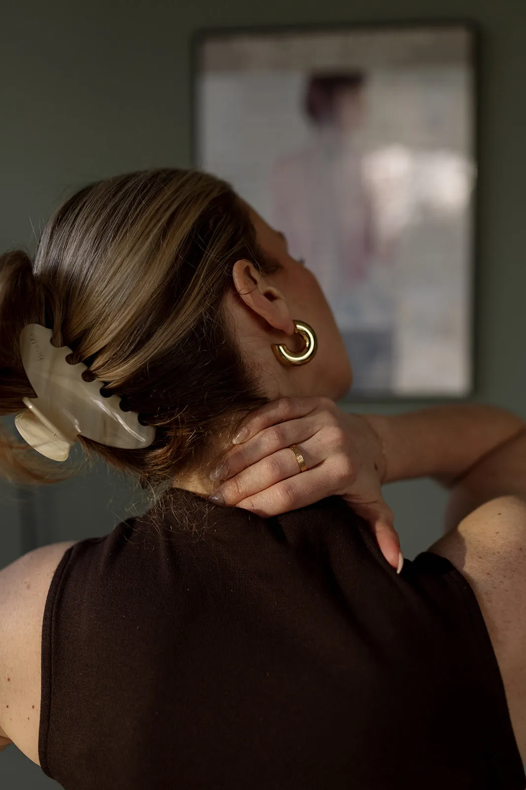 Woman with light brown hair in a clip, wearing gold hoop earring and gold ring, touching her neck.