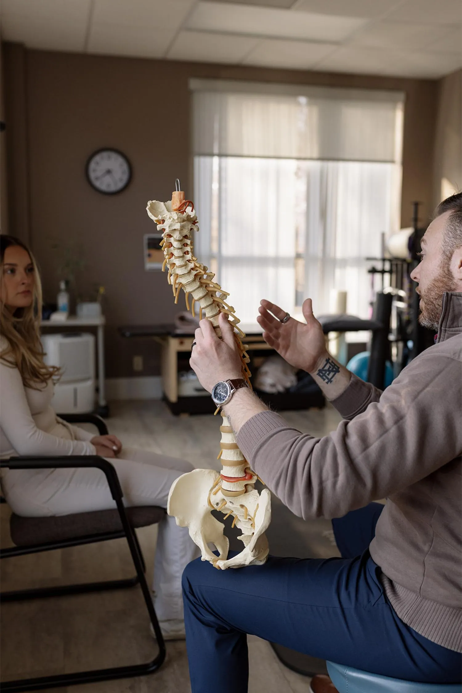 Man explaining spine anatomy with vertebrae model to woman seated in a medical office.