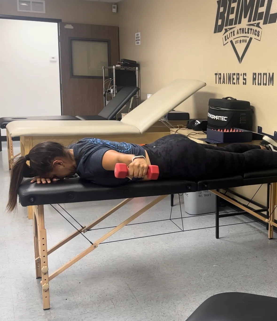 Woman lying face down on a massage table holding a pink dumbbell, exercising in a trainer's room.
