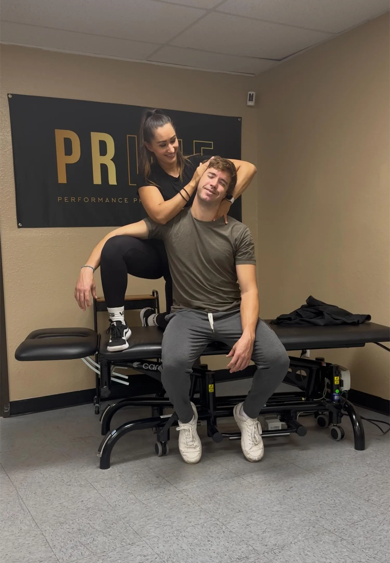 Woman in black outfit stretching a man's neck while he sits on a therapy table in a clinic.