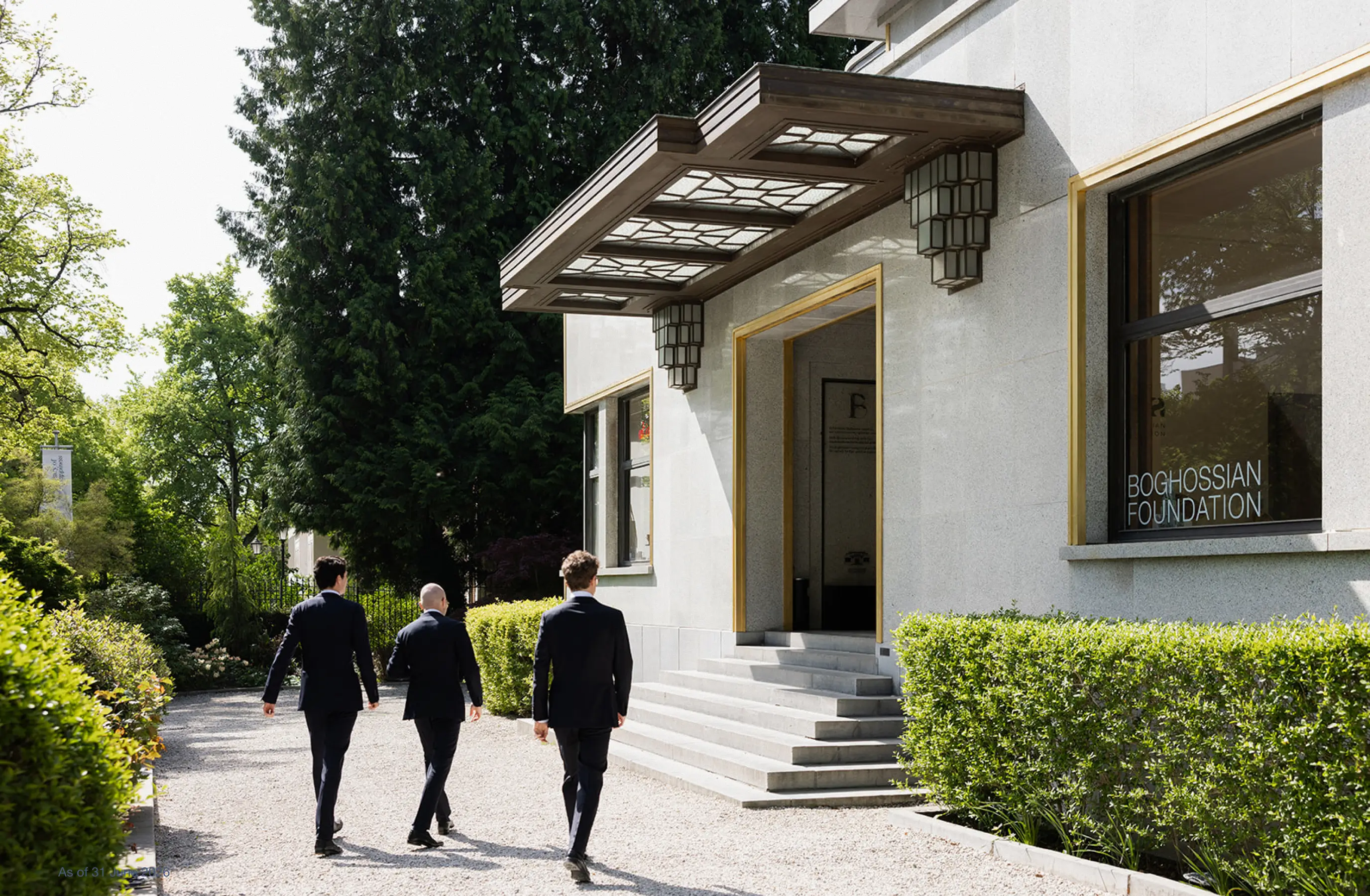 Three men in suits walk on a gravel path toward the entrance of the Boghossian Foundation building.