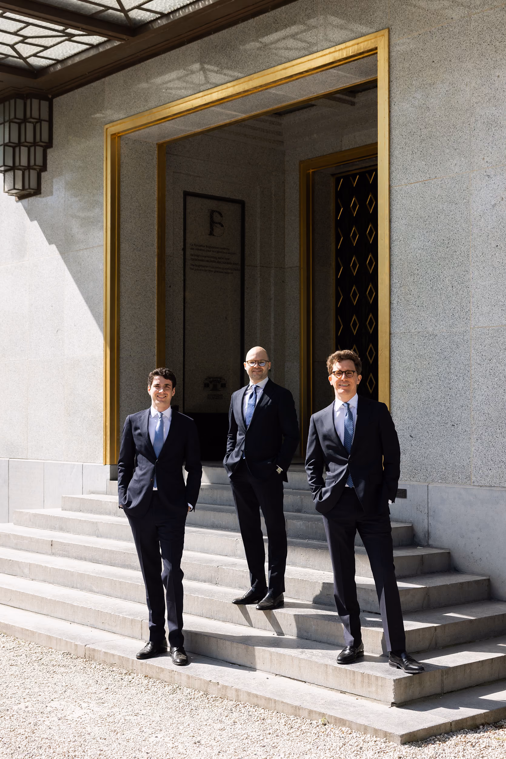 Three men in dark suits standing on stone steps outside a building with a gold-framed entrance.