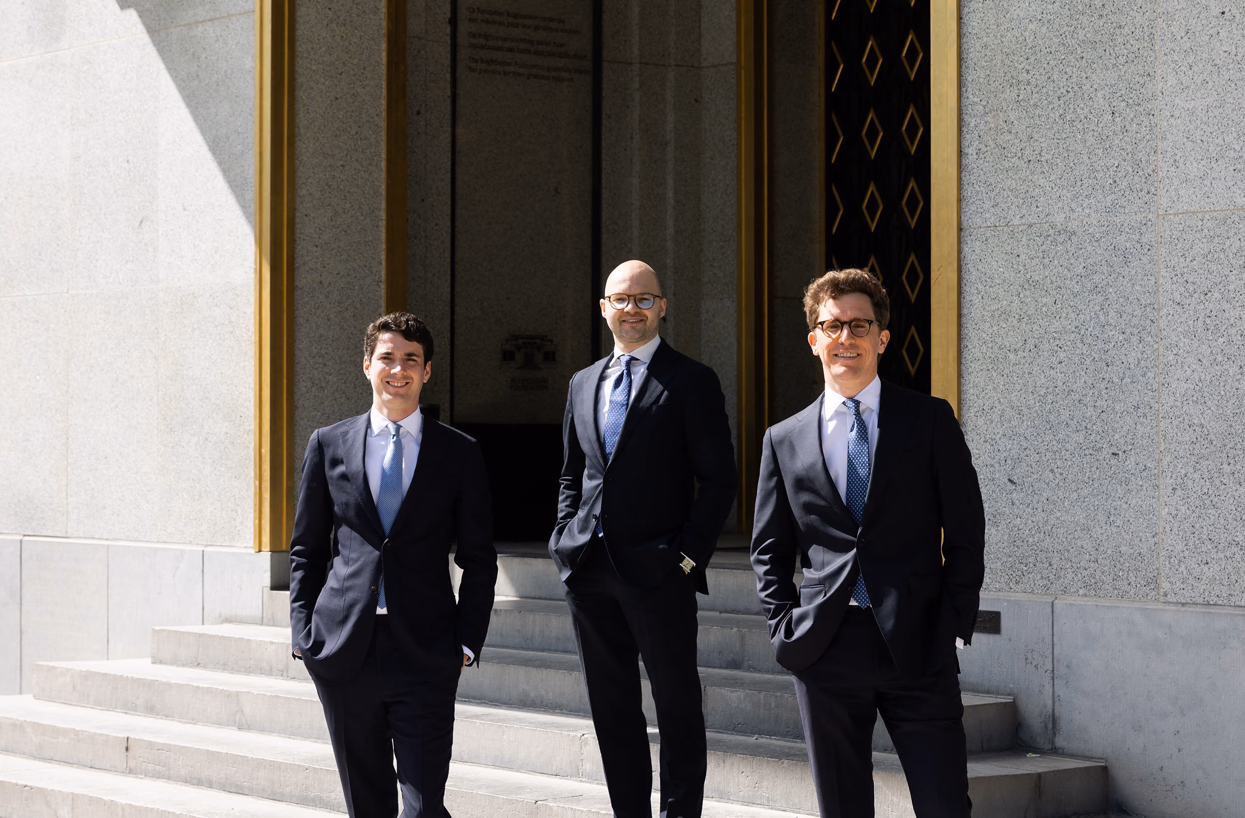 Three men in suits standing on concrete steps in front of a building with gold-trimmed doors.