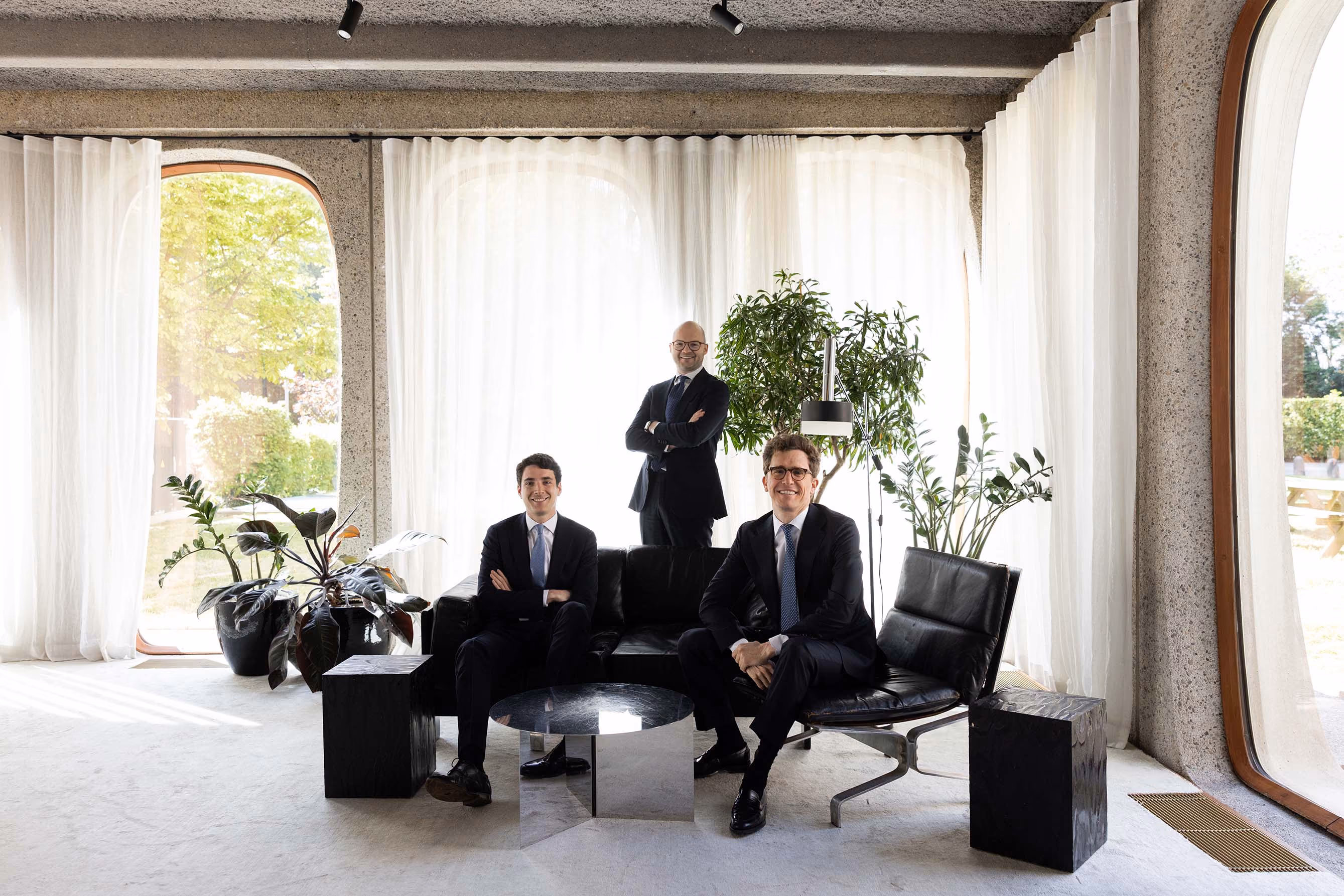 Three men in suits posing in a bright room with large windows, plants, and modern furniture.