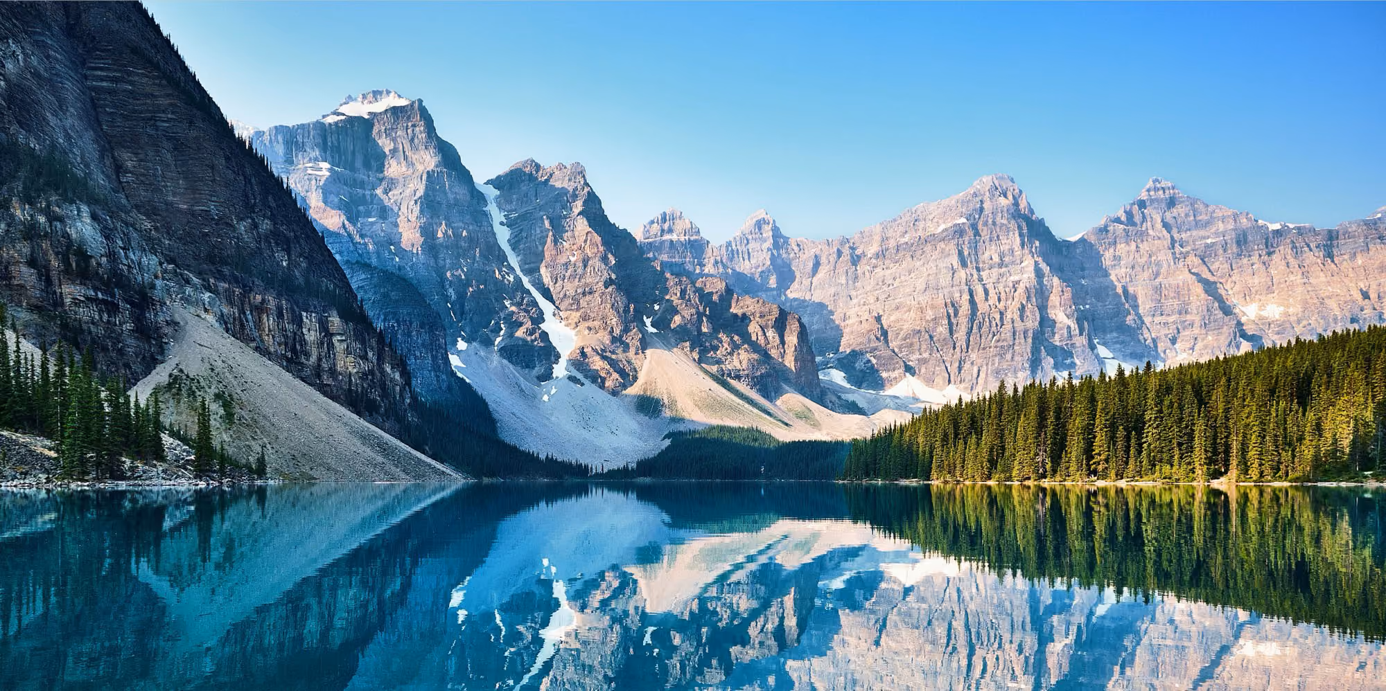 Mountain peaks reflected in a clear blue lake bordered by dense pine forest under a clear sky.