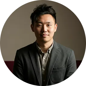 Portrait of a young Asian man with short black hair wearing a dark blazer and light shirt against a muted background.