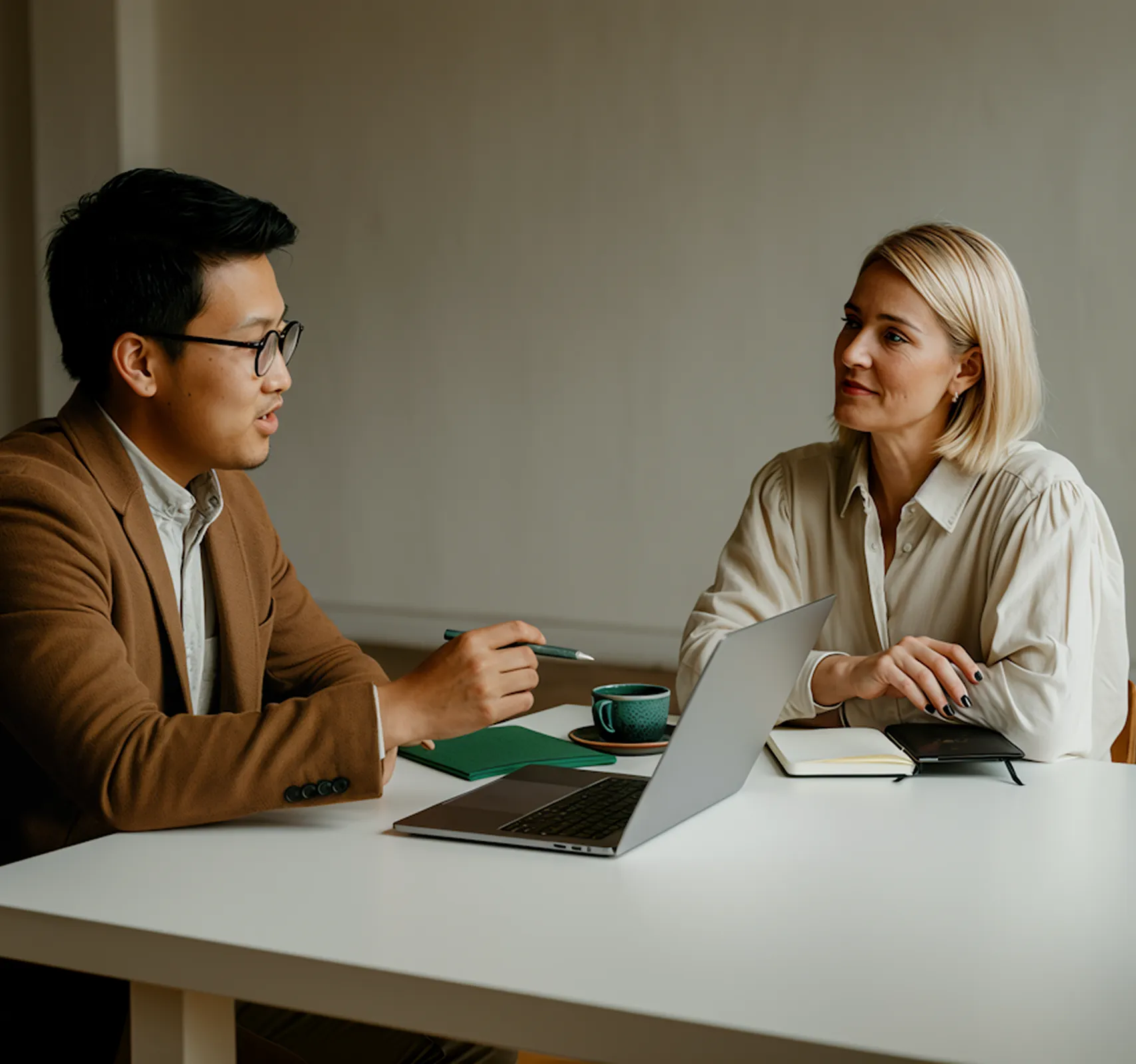 A man in a brown blazer and glasses talks to a woman with blonde hair seated across a table with a laptop and notebooks.