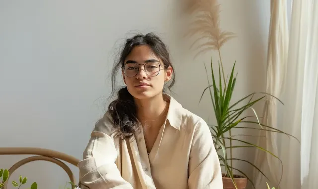 Young woman with glasses and loose hair wearing a beige shirt sitting indoors with plants in the background.