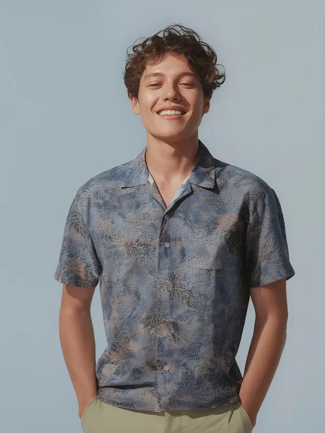 Smiling young man with curly hair wearing a patterned short-sleeve shirt and light pants against a plain blue background.