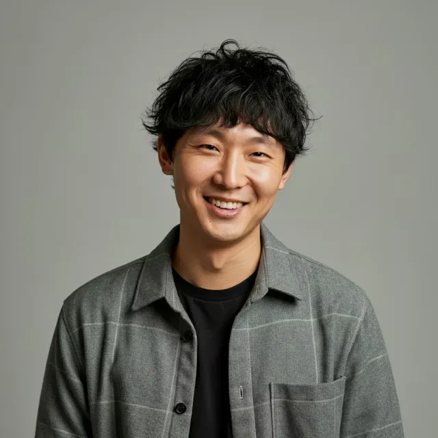 Smiling young man with curly black hair wearing a grey checkered shirt over a black t-shirt against a plain grey background.