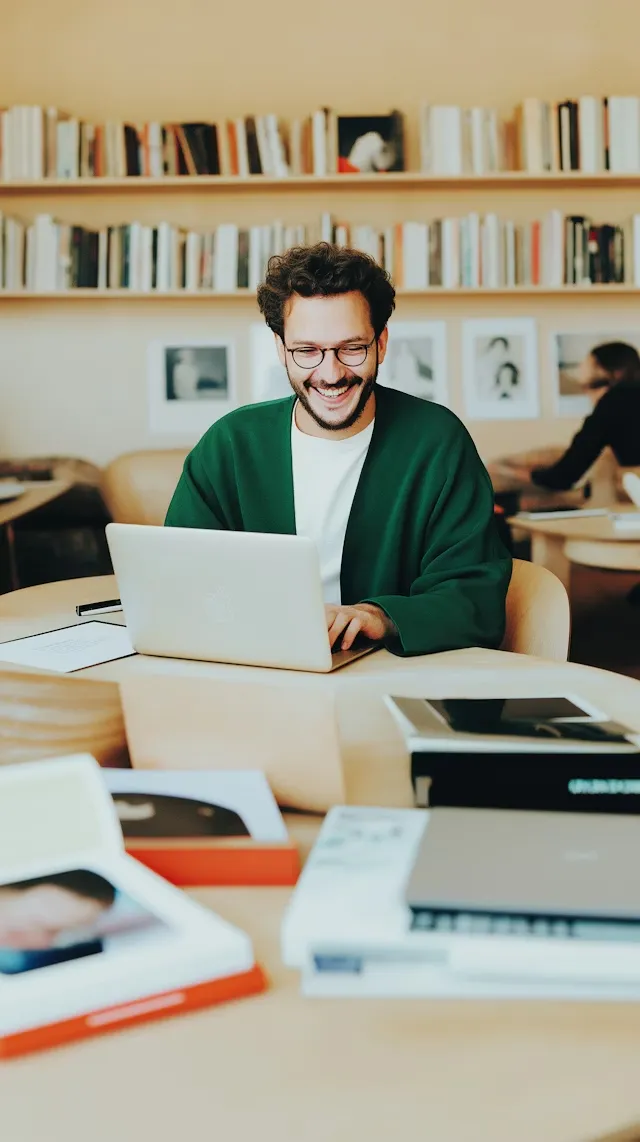 Smiling man wearing glasses and a green cardigan working on a laptop in a cozy library with bookshelves in the background.