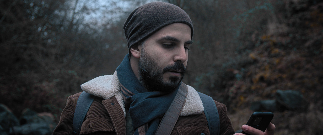 Man wearing a beanie and scarf outdoors looking at his smartphone in a wooded area.