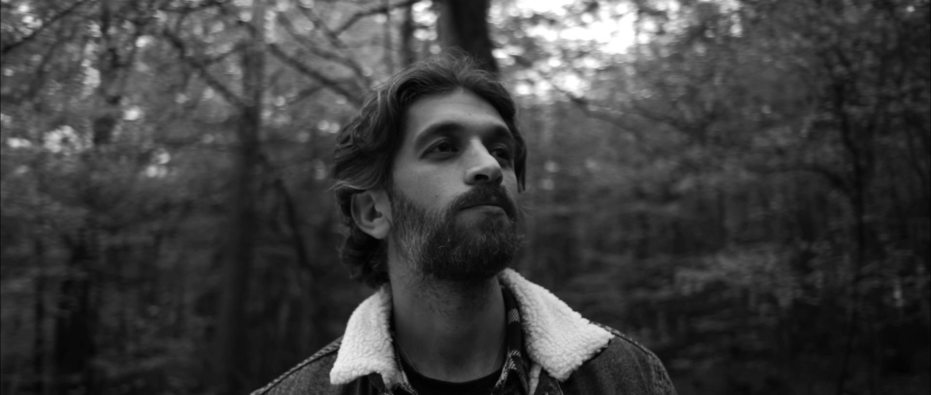 Black and white photo of a bearded man in a shearling jacket looking upward in a forest.