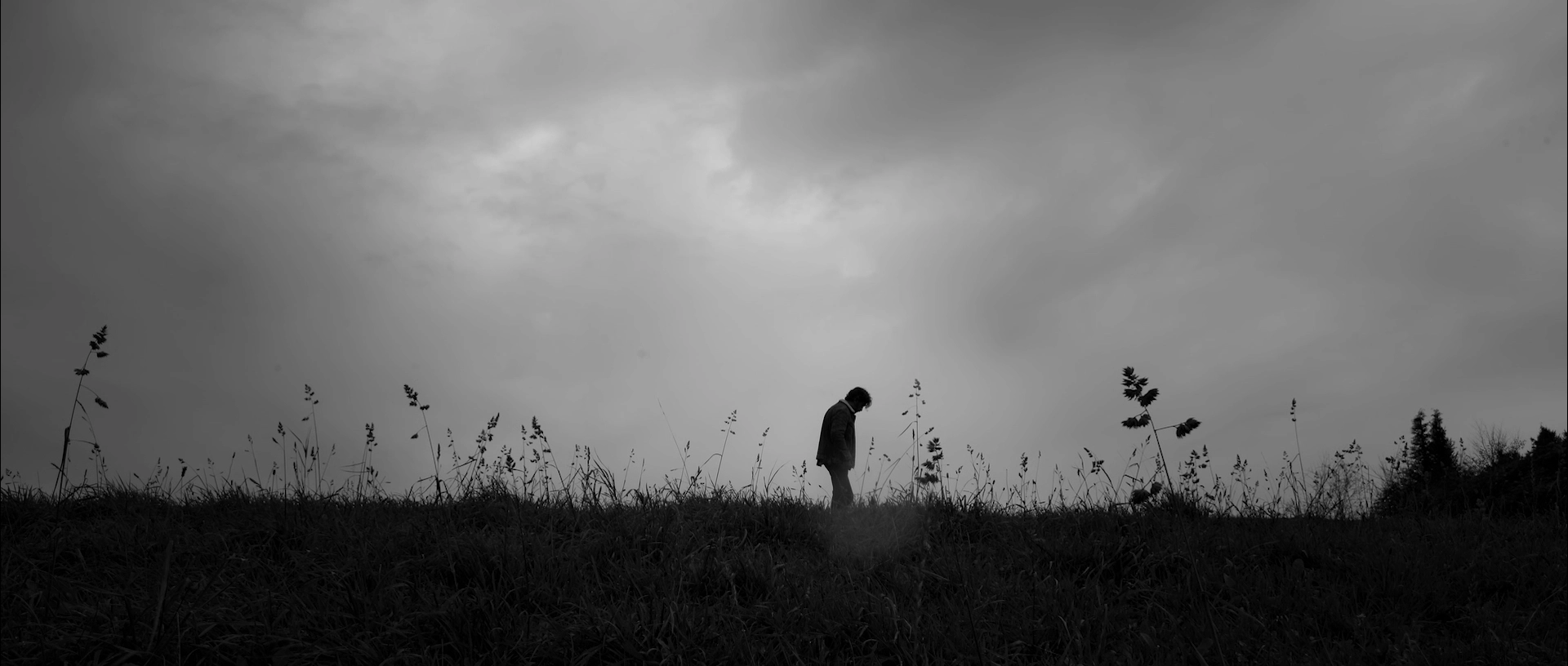 Silhouette of a person standing alone on grassy hill under a cloudy sky in black and white