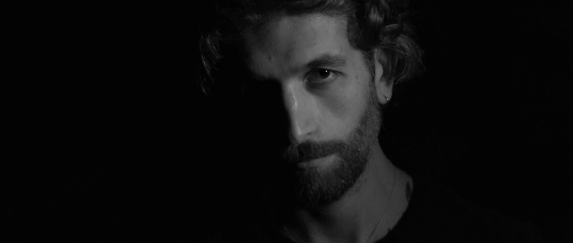 Black and white close-up of a man with beard, mustache, and earring, half his face in shadow.