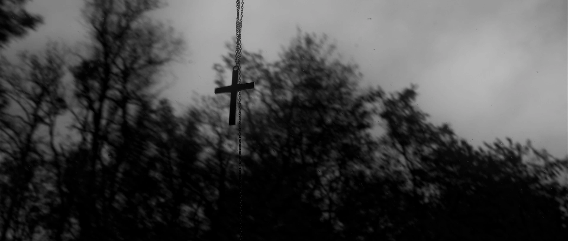 Black cross pendant hanging on a chain in front of blurred dark trees and cloudy sky.