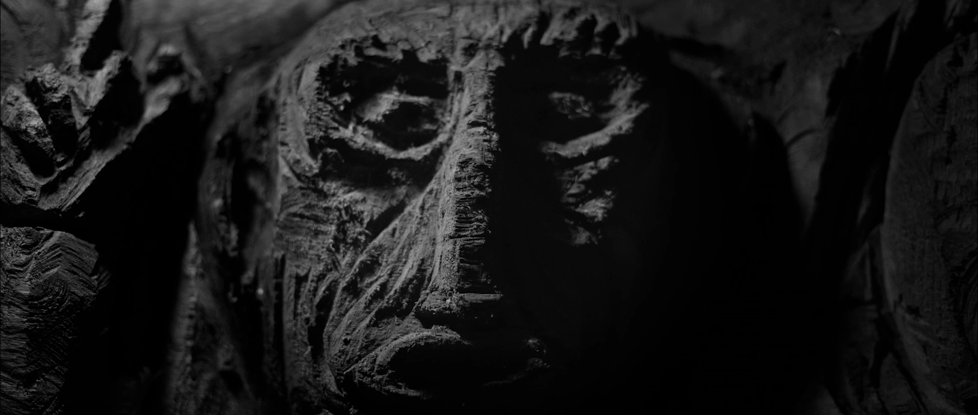 Close-up of a dark, textured wooden carving depicting a somber face in deep shadows.