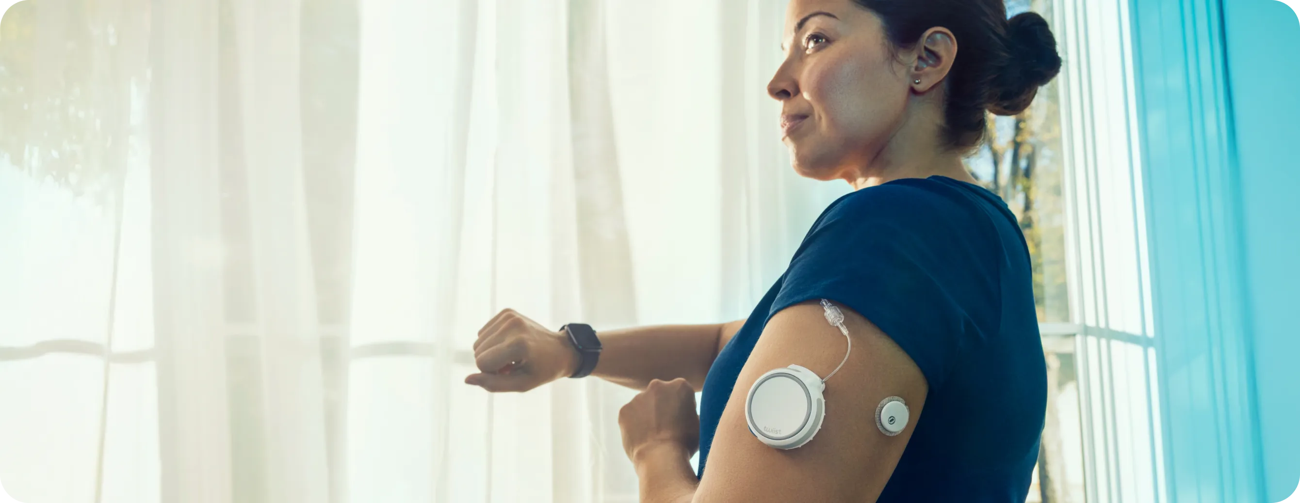 Woman with hair bun wearing a blue shirt and insulin pump devices on upper arm checking smartwatch.