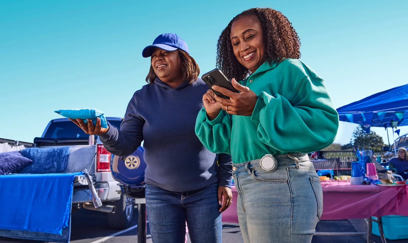 Two smiling women at a tailgate, one holding a cornhole bag and one checking her phone while wearing the twiist insulin pump on her waistband.