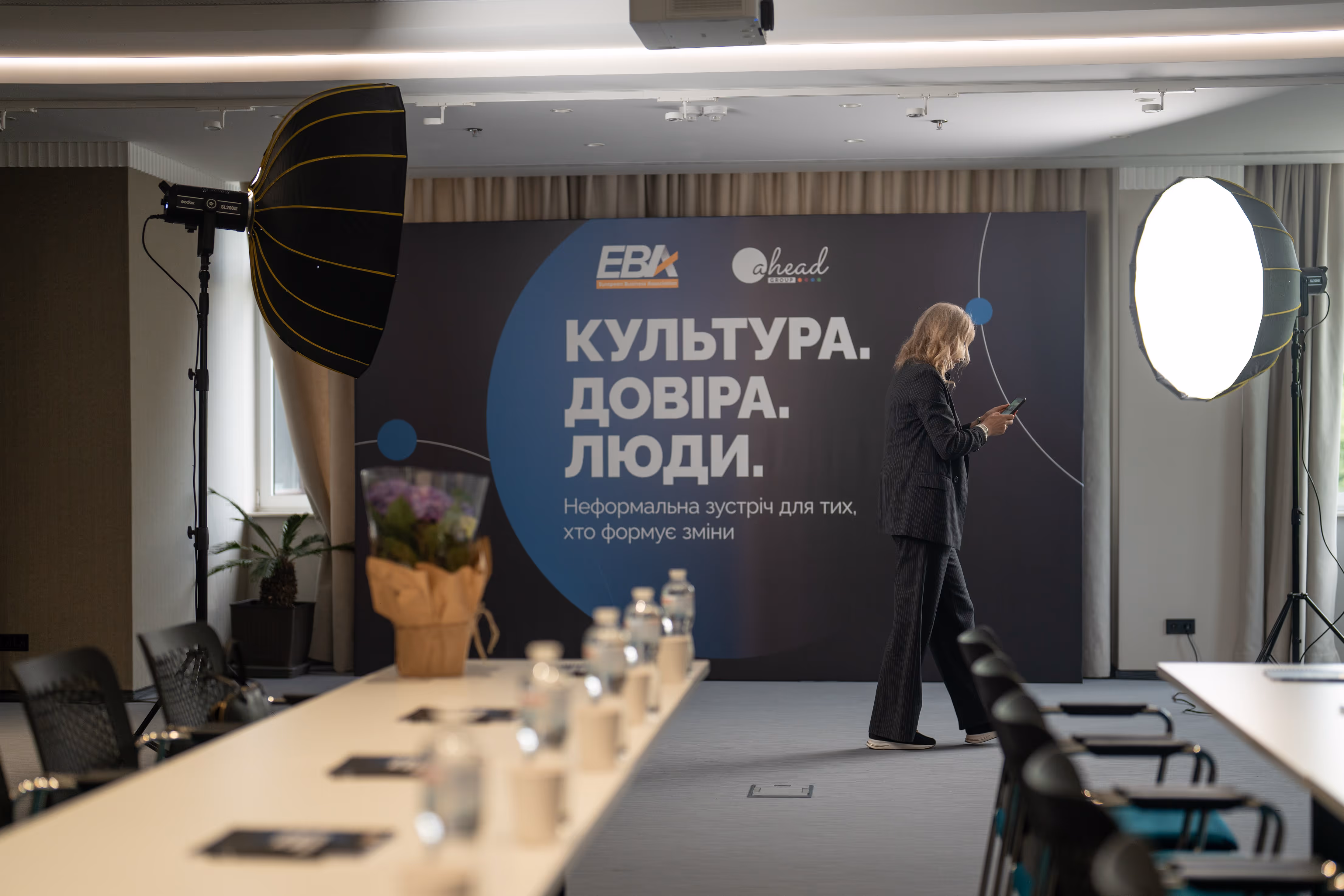 Жінка в костюмі стоїть біля банера з текстом про культуру, довіру та людей у конференцзалі.