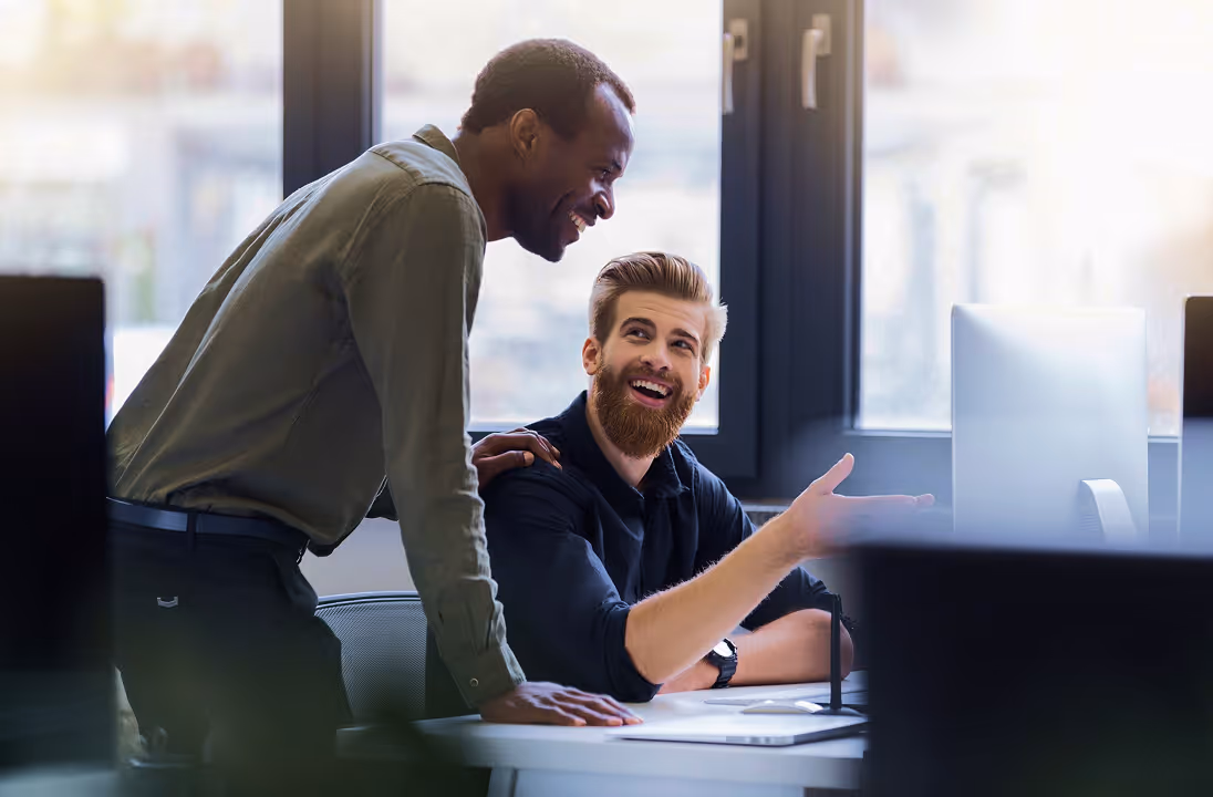 Two male colleagues smiling and talking at a desk with a computer in a bright office.