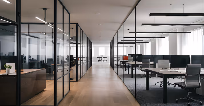 Modern office hallway with glass-walled meeting rooms on the left and open office desks with chairs on the right.
