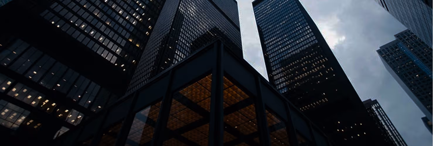 Low-angle view of tall office buildings with glass windows against a cloudy sky at dusk.