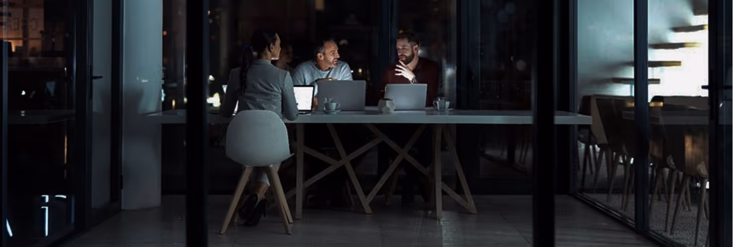 Three people with laptops engaged in a nighttime meeting in a modern glass-walled office.