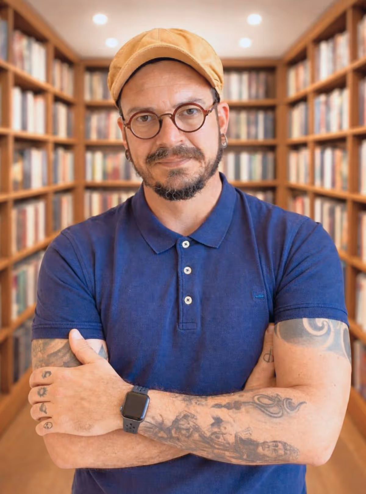 Homem de camiseta azul e boné bege com braços cruzados e tatuagens em uma biblioteca.