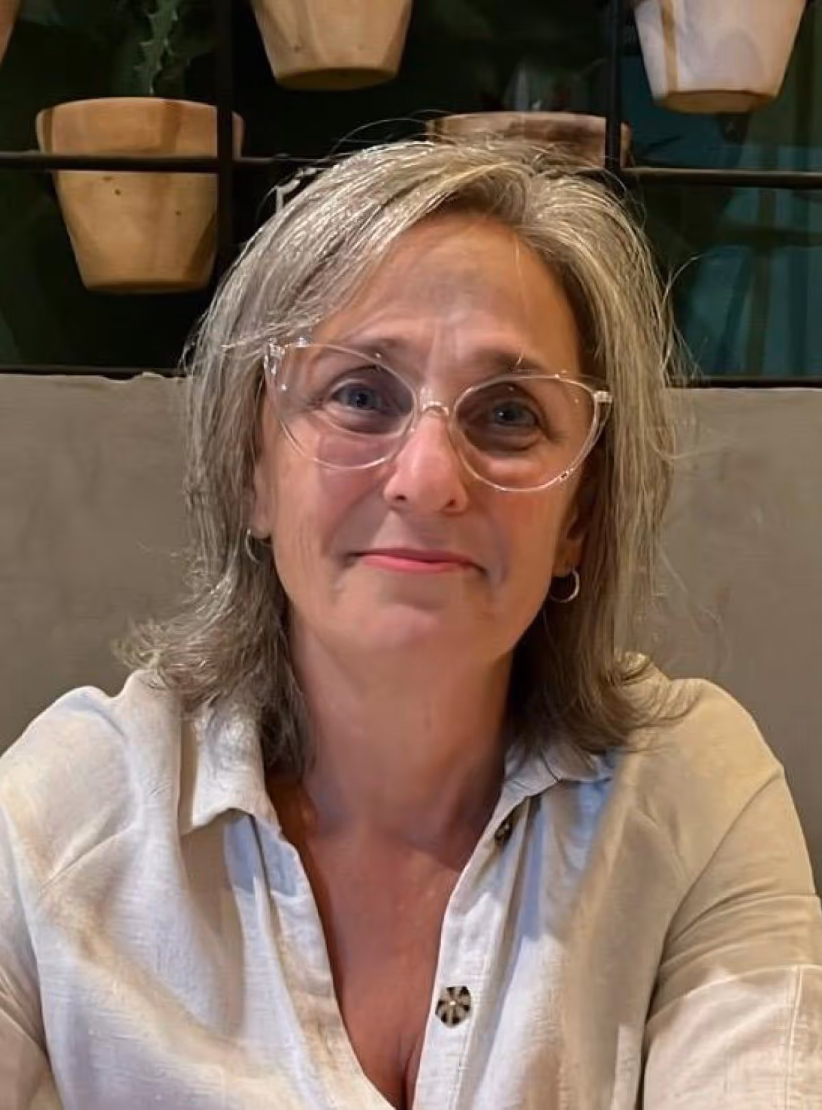 Mulher de cabelos grisalhos e óculos transparentes sorrindo, vestindo camisa branca.