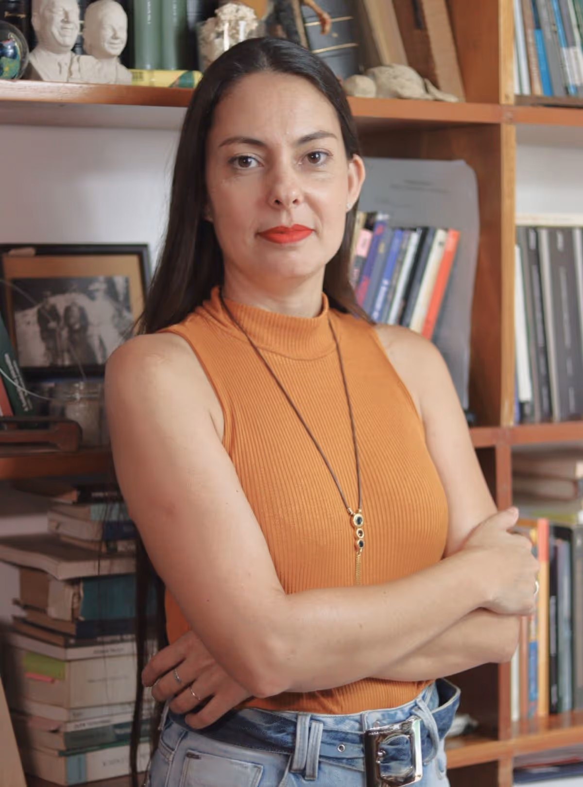 Mulher de blusa sem mangas laranja e calça jeans azul posa com braços cruzados em biblioteca.
