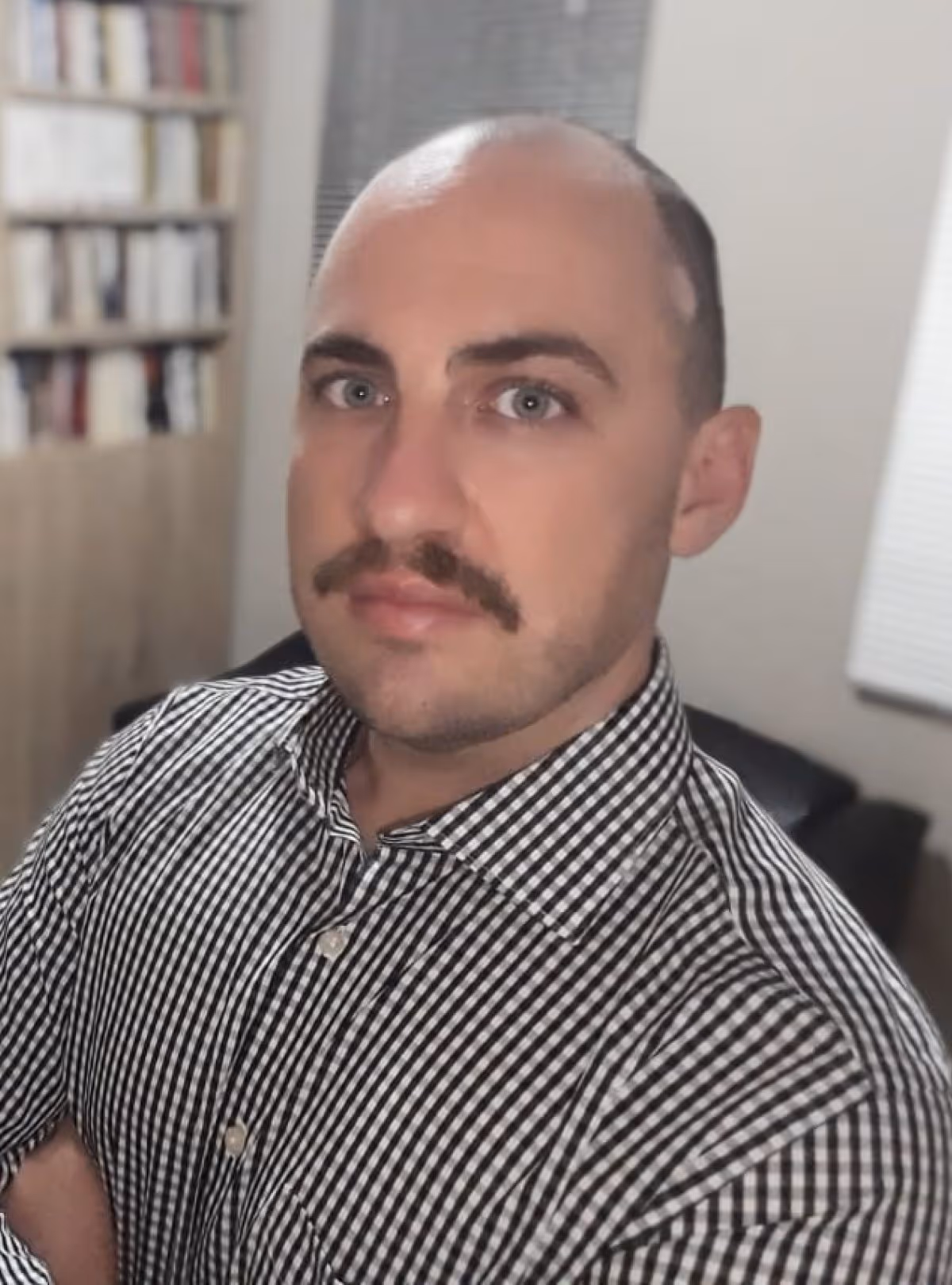 Homem com bigode e camisa xadrez preta e branca em ambiente interno com estante e janela ao fundo.