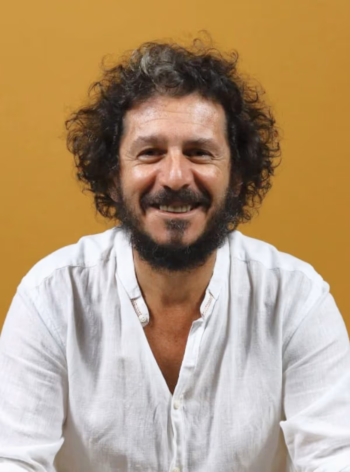 Homem sorridente com cabelo e barba cacheados, vestindo camisa branca, fundo amarelo.