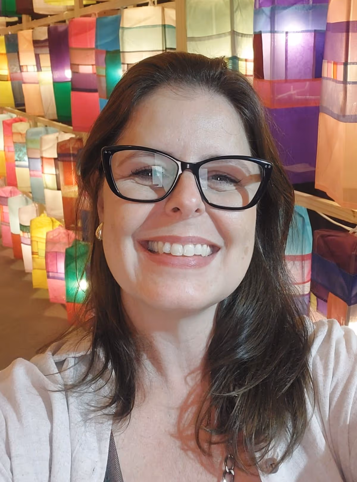 Mulher sorridente usando óculos pretos com fundo de lanternas coloridas iluminadas.