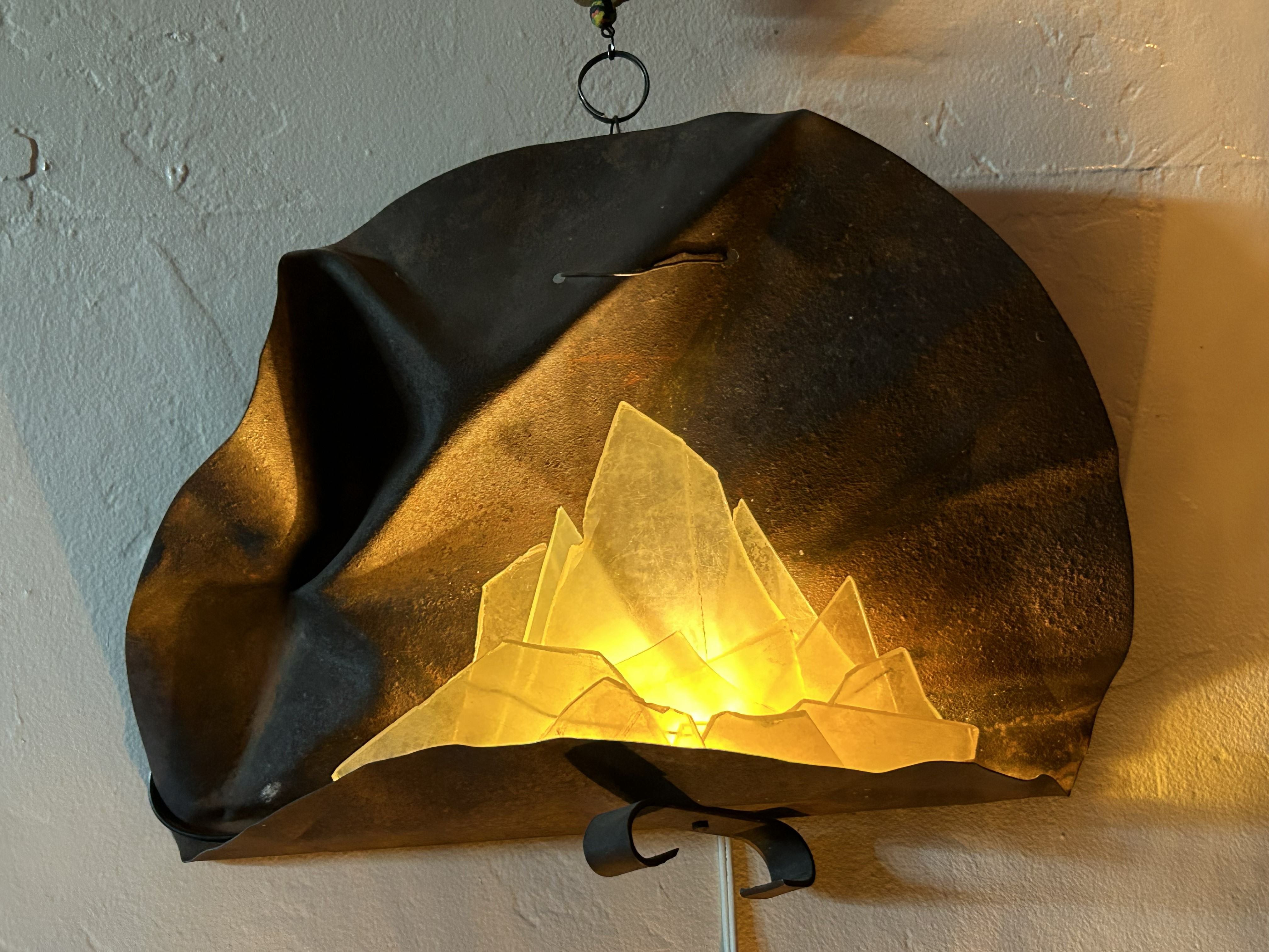 Wall-mounted lamp with jagged glass pieces glowing warmly inside a crumpled metal shade.