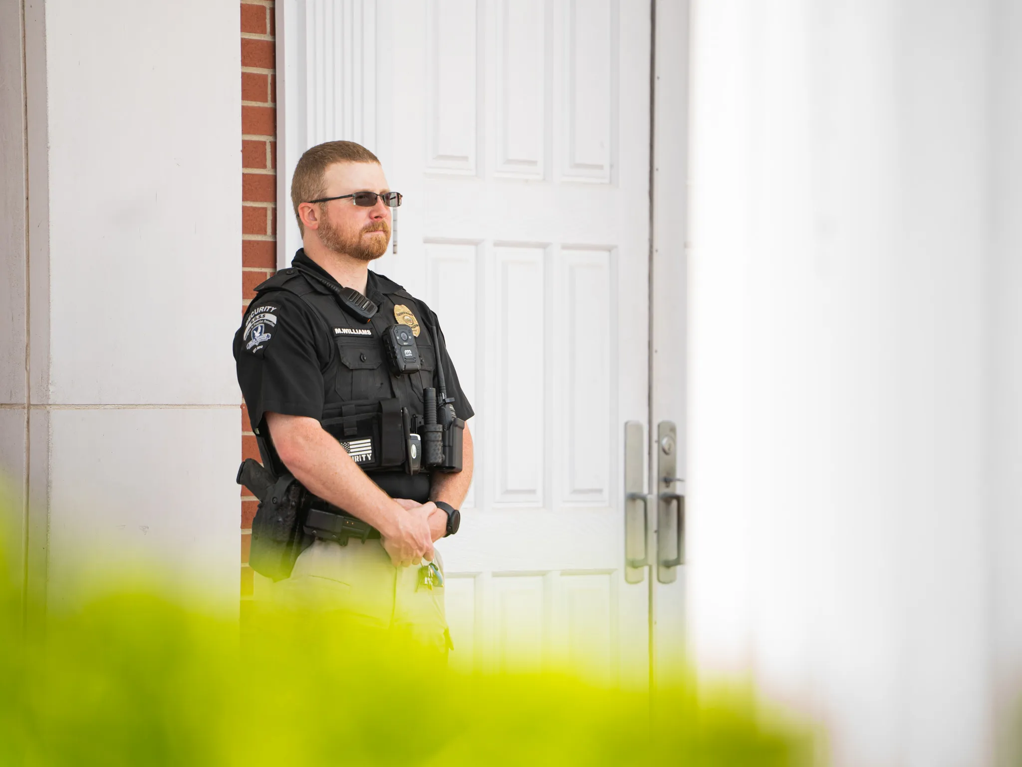Security officer with beard and glasses standing guard by white double doors with brick trim.