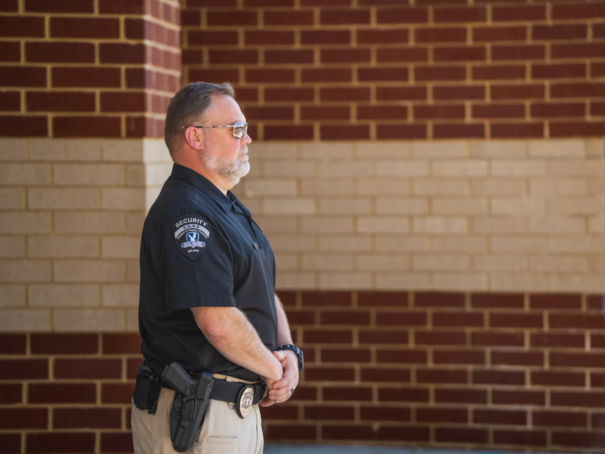 Security guard in uniform and sunglasses standing in front of a brick wall with hands clasped.