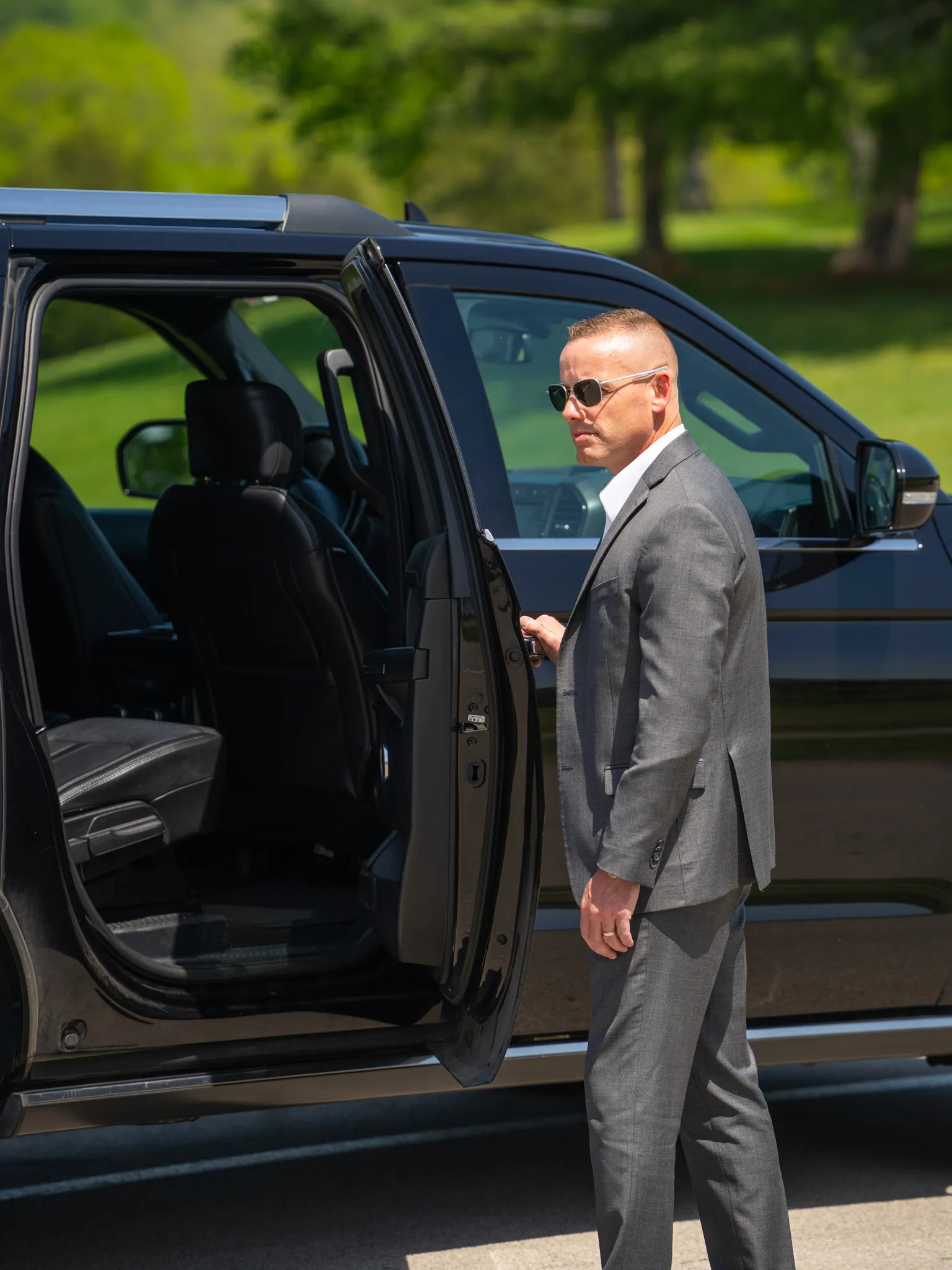 Man in a gray suit and sunglasses opening the rear door of a black SUV outdoors.