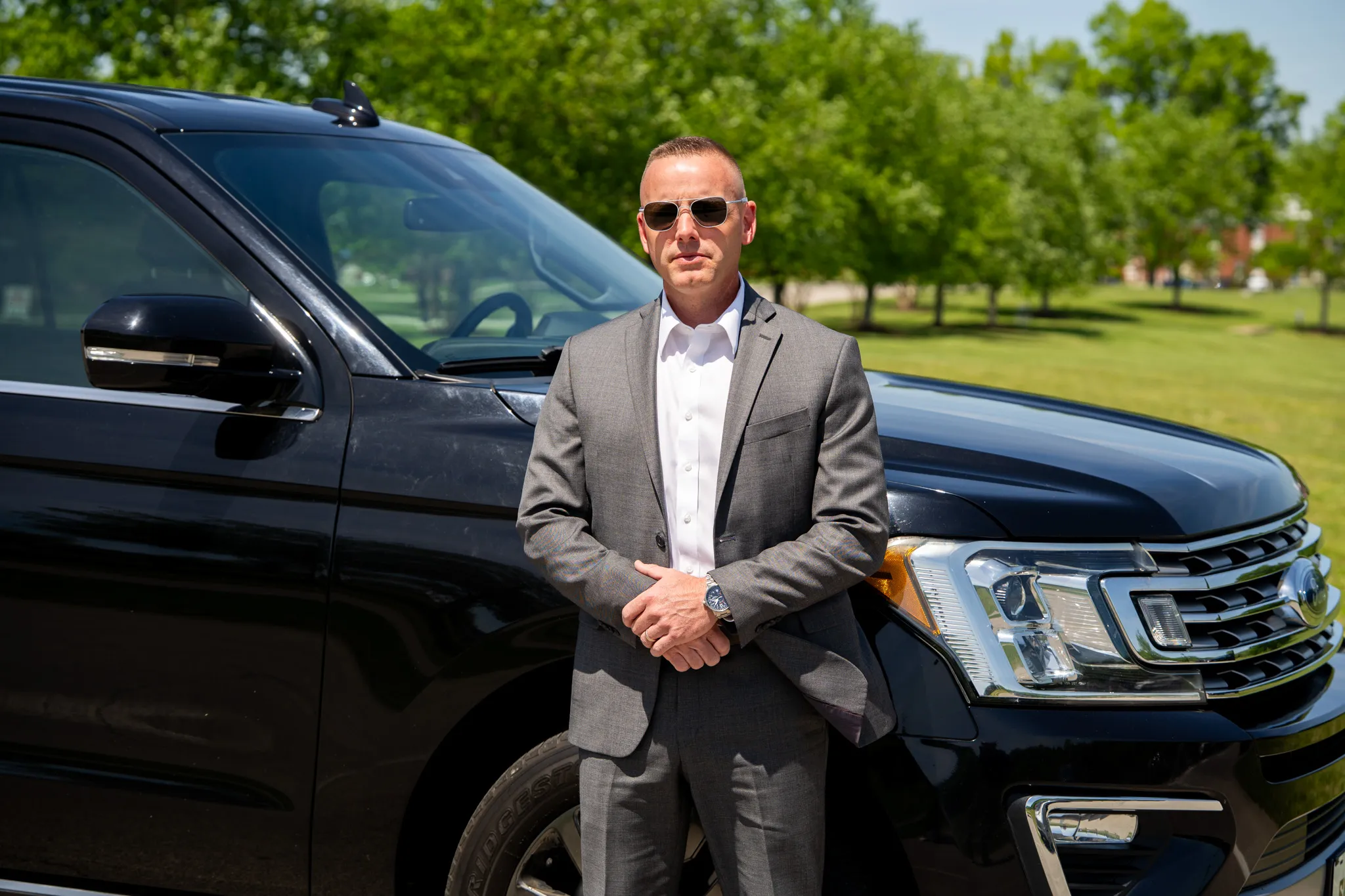 Man in a gray suit and sunglasses stands in front of a black SUV outdoors on a sunny day.