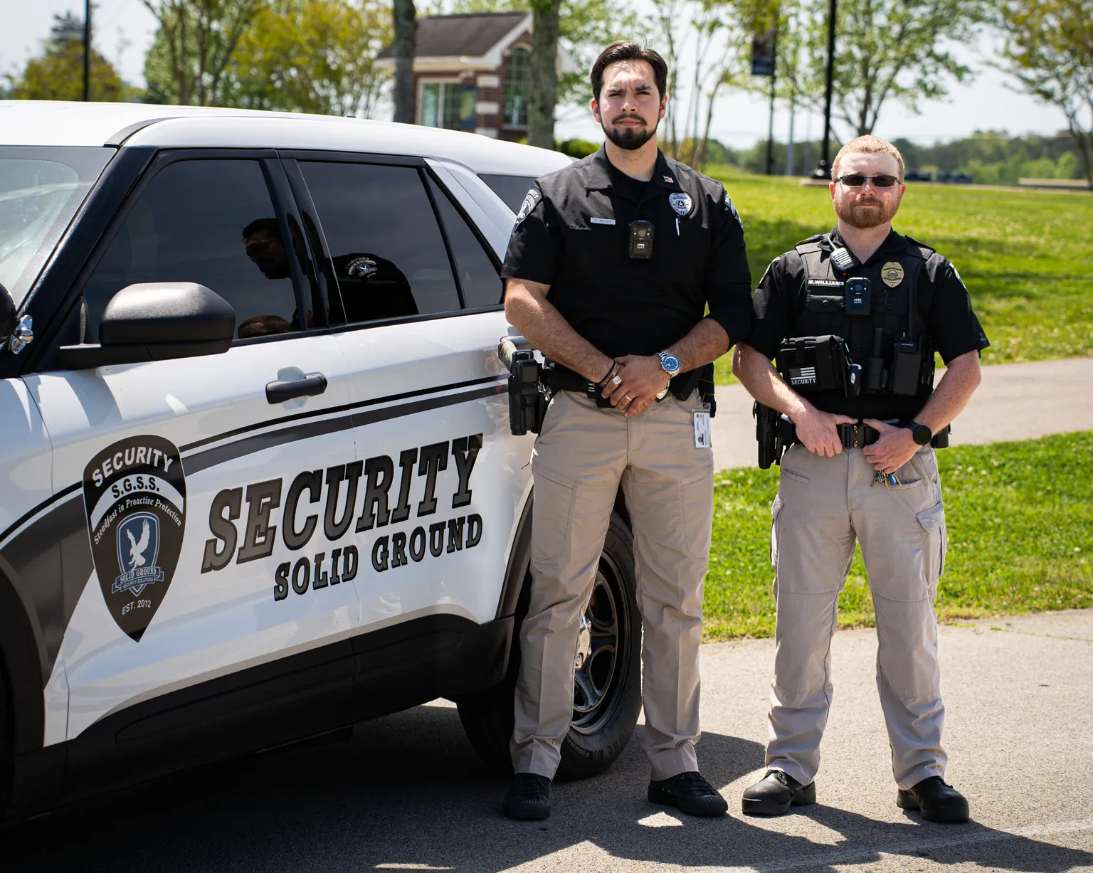 Two security officers standing by a white patrol vehicle labeled Security Solid Ground on a sunny day.