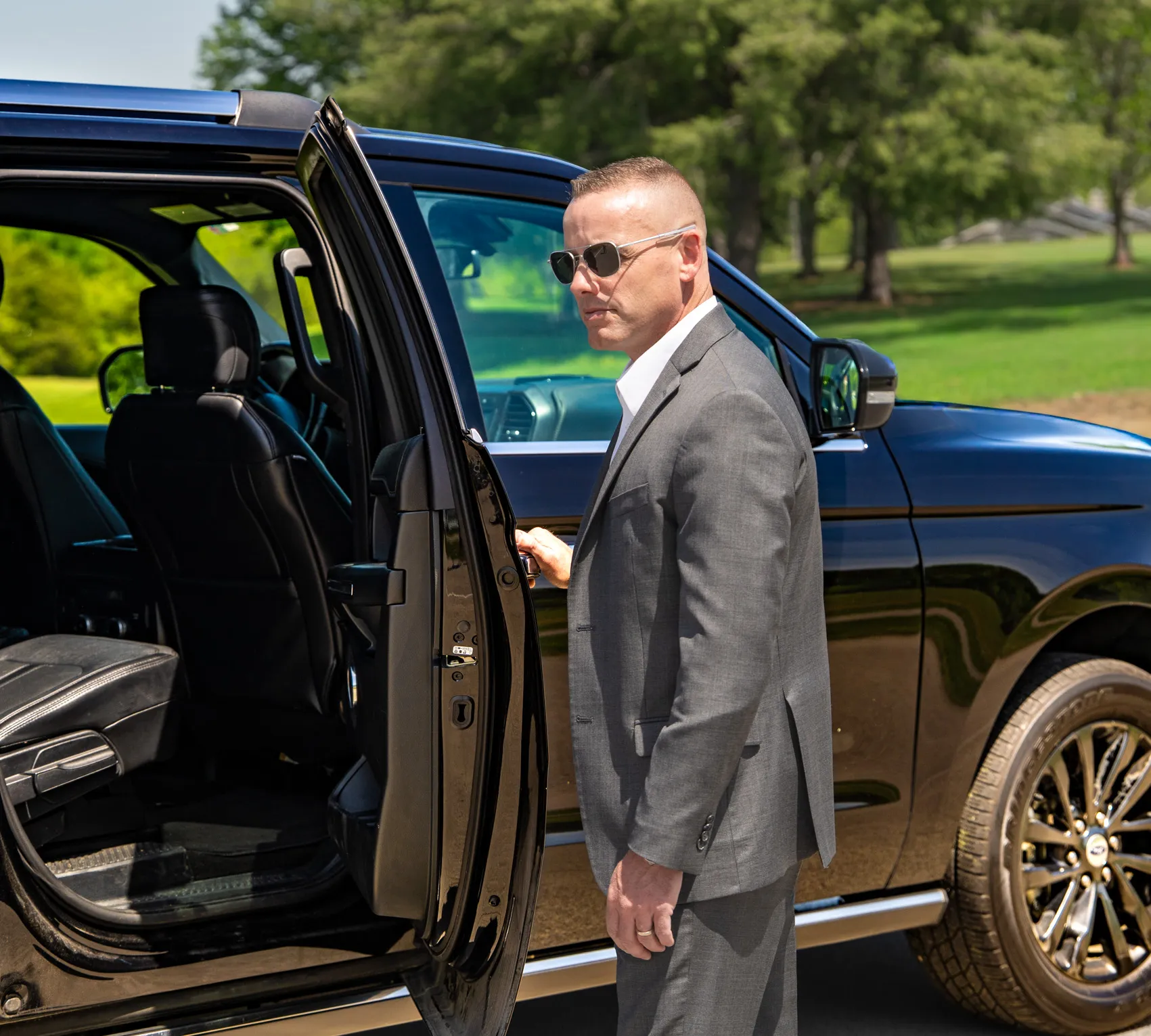 Man in gray suit and sunglasses opening rear door of black SUV in a green outdoor setting.