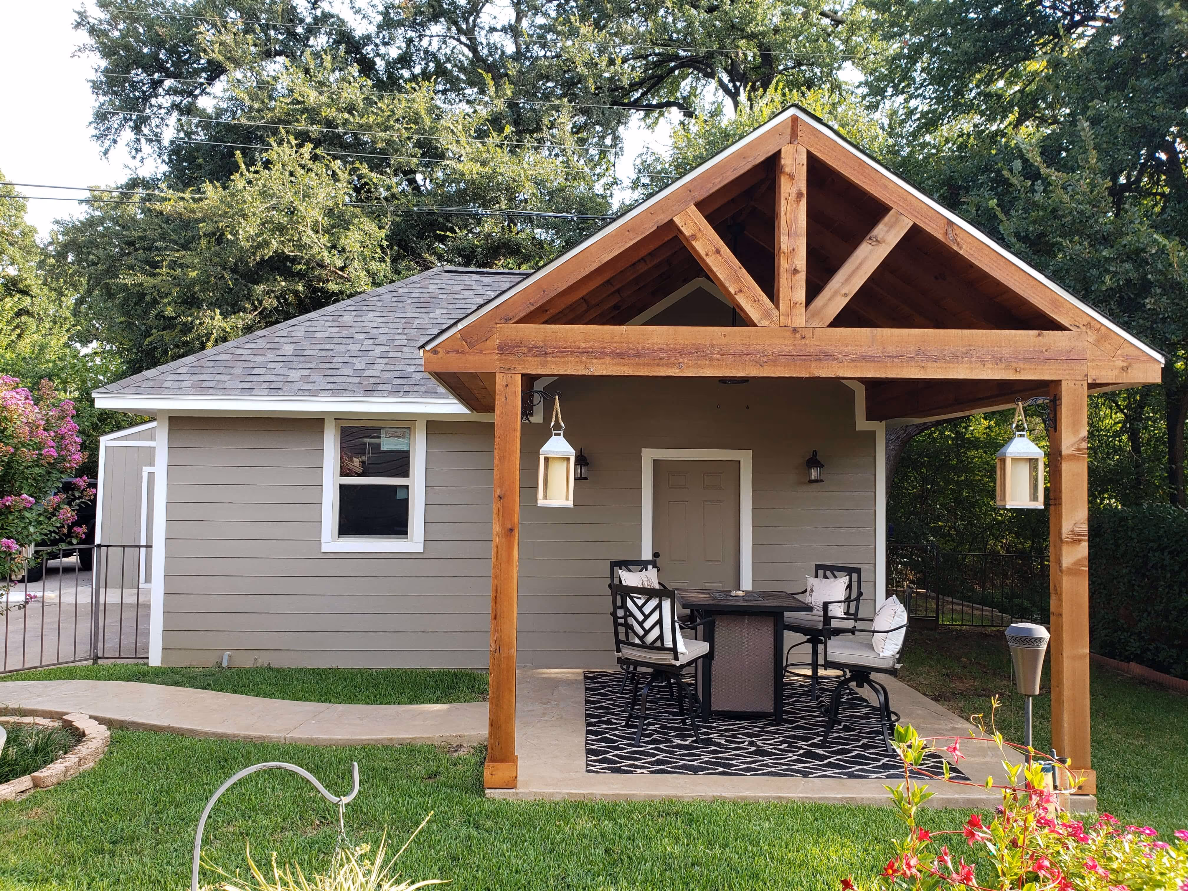 Back porch with wooden pergola, patio table, four chairs, hanging lanterns, and a black patterned rug.