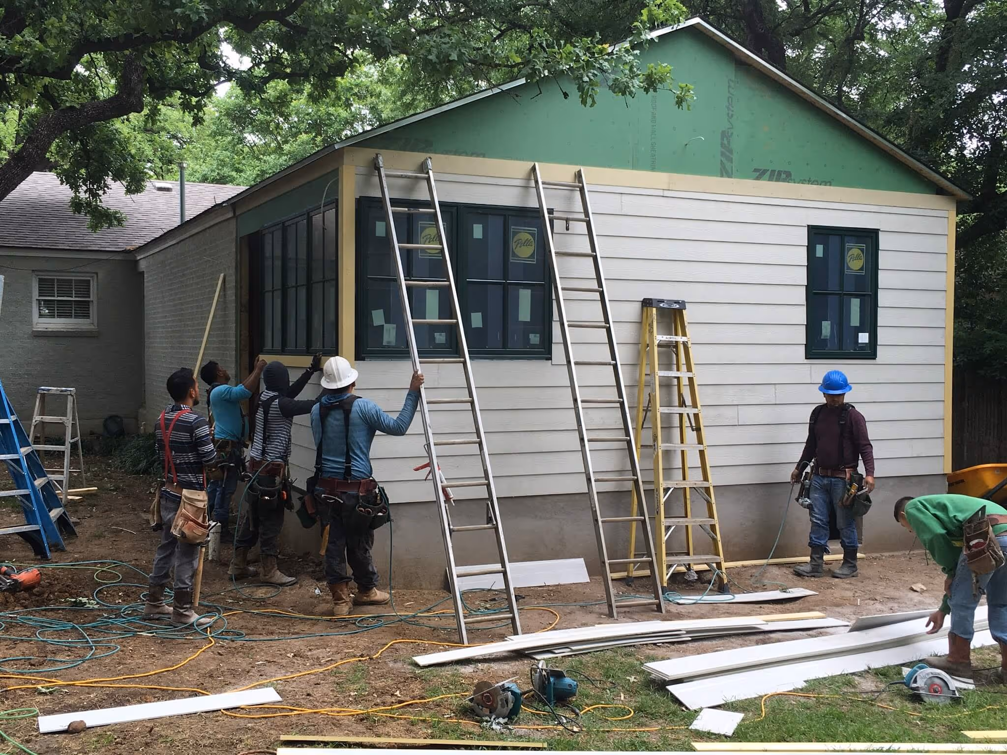 Construction workers installing siding on house exterior with ladders and tools around the site.